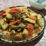 A vibrant plate of Moroccan couscous topped with colorful vegetables, chickpeas, and tender pieces of lamb, served with a side of spicy harissa sauce.
