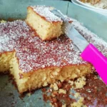 A homemade coconut cake with layers of creamy frosting and a generous sprinkle of shredded coconut, served on a rustic wooden table.